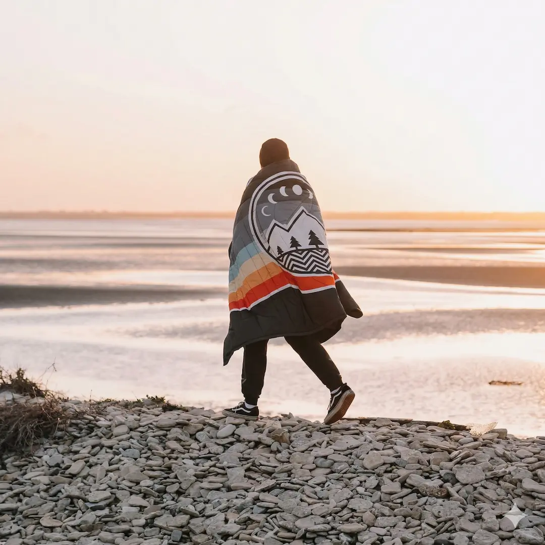 Man walking along a pebbly beach wrapped in a VOITED Ripstop Camping Blanket